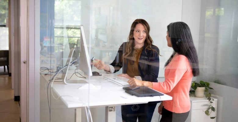 Two women talking at a standing desk in a glass-walled office workspace